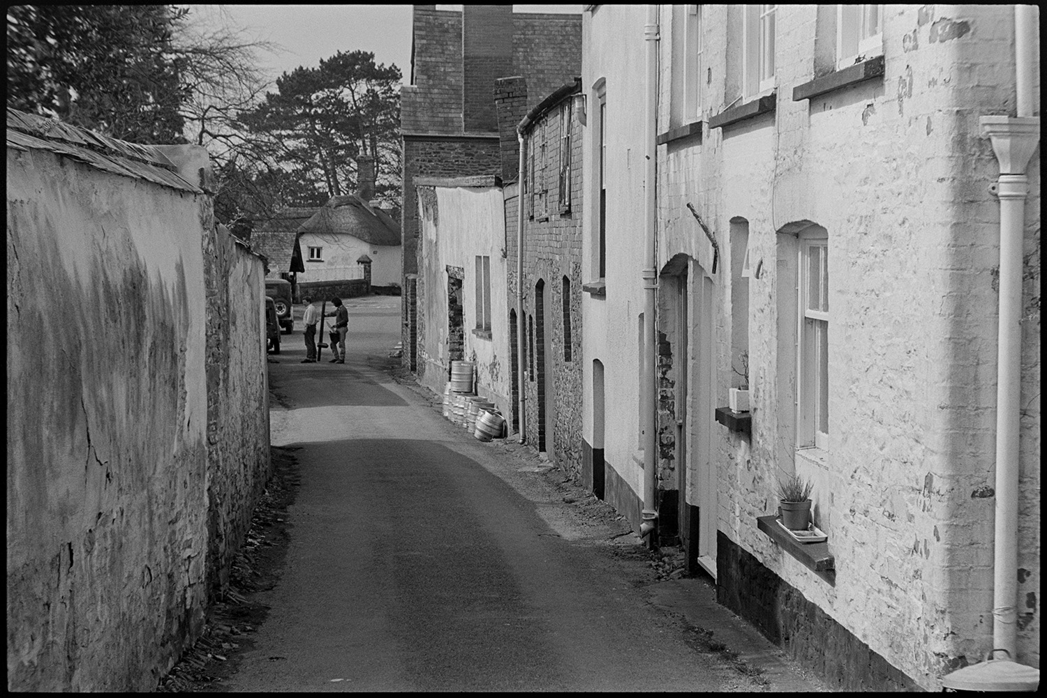 Street scene with parked motor bikes.
[A narrow street near the church and the old school at Chulmleigh. There are metal beer barrels stacked outside a property. Two young men and parked vehicles are at the end of the street, with a thatched house, railings, and tall fir trees in the background.]