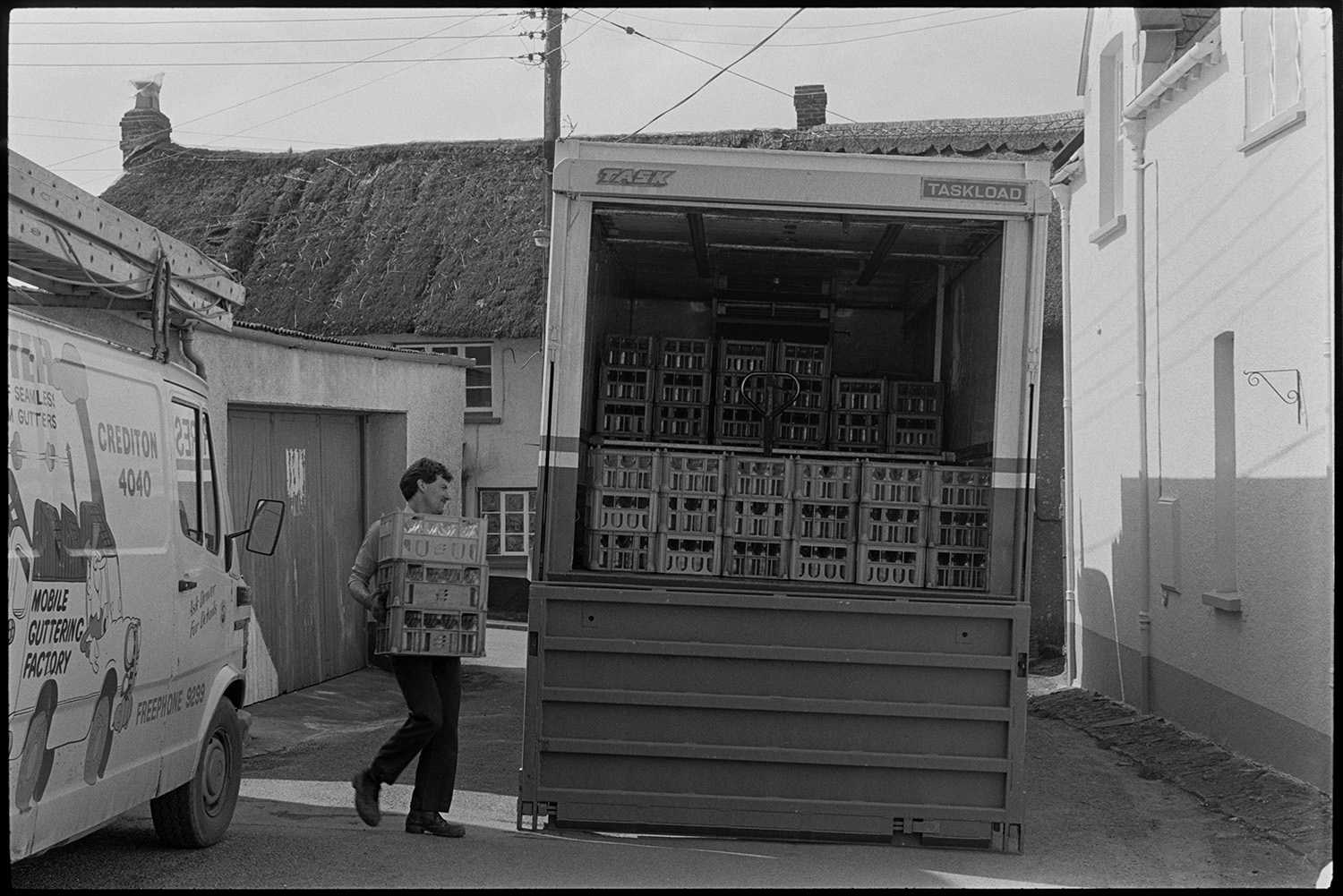 Lorry unloading milk crates.
[A man unloading or loading crates with milk bottles from an Express Dairy lorry parked in New Street, Chulmleigh. There is a van belonging to a gutter repair company parked alongside the lorry, and a row of thatched cottages in the background.]