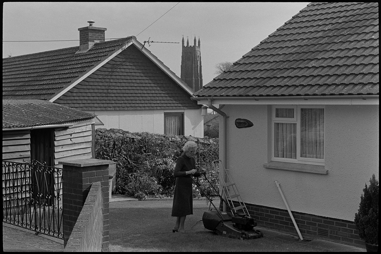 Street scenes, public bench.
[A woman mowing her lawn in the garden of a bungalow in Chulmleigh. There is a garden shed, wrought iron gates and garden plants in front of a wall. Another bungalow is next door, with the tower of Chulmleigh church in the background.]