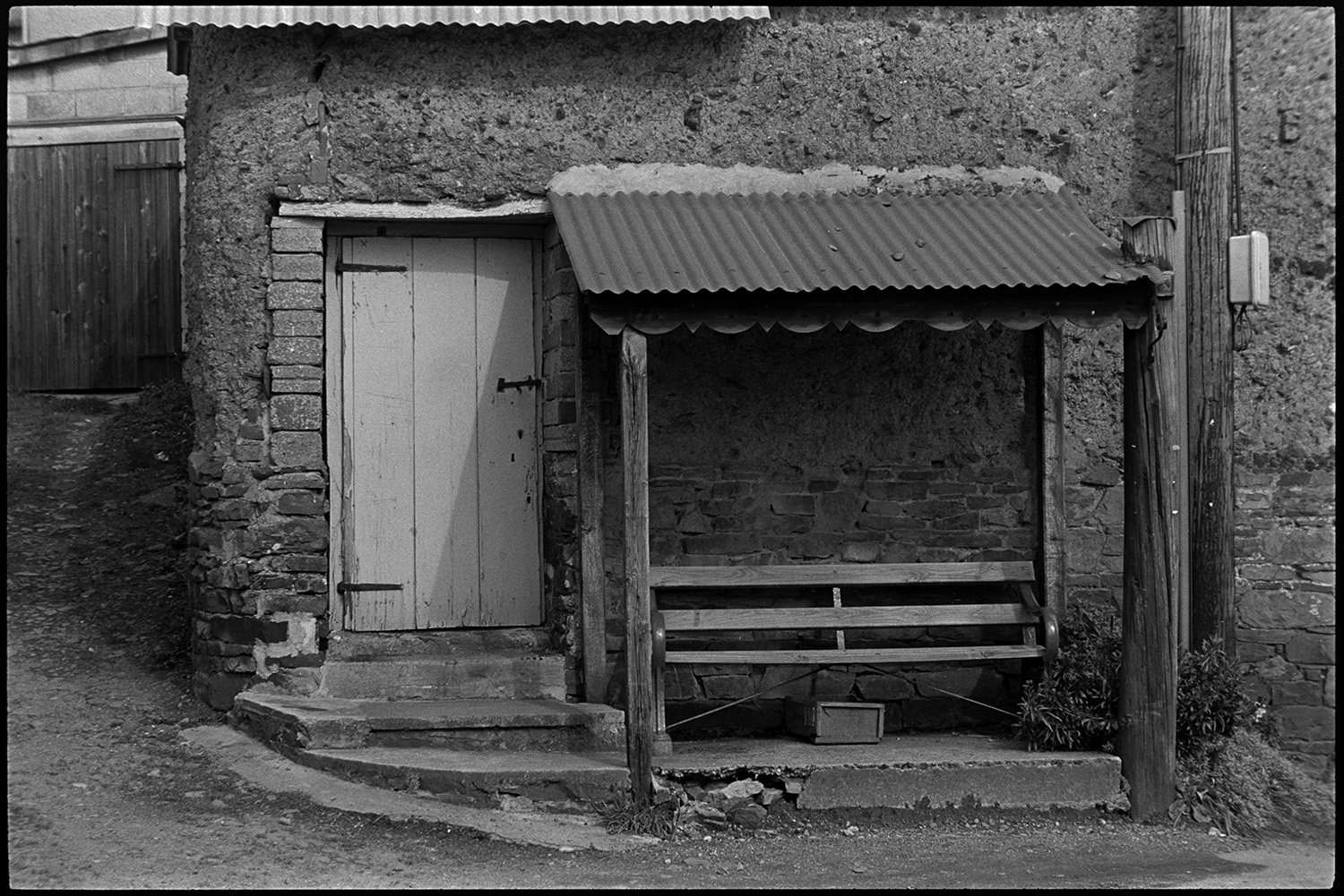 Street scenes, public bench.
[A public bench with wooden supports and a corrugated iron roof on a street corner in Chulmleigh. The shelter is attached to a cob barn with steps leading up to a wooden door.]