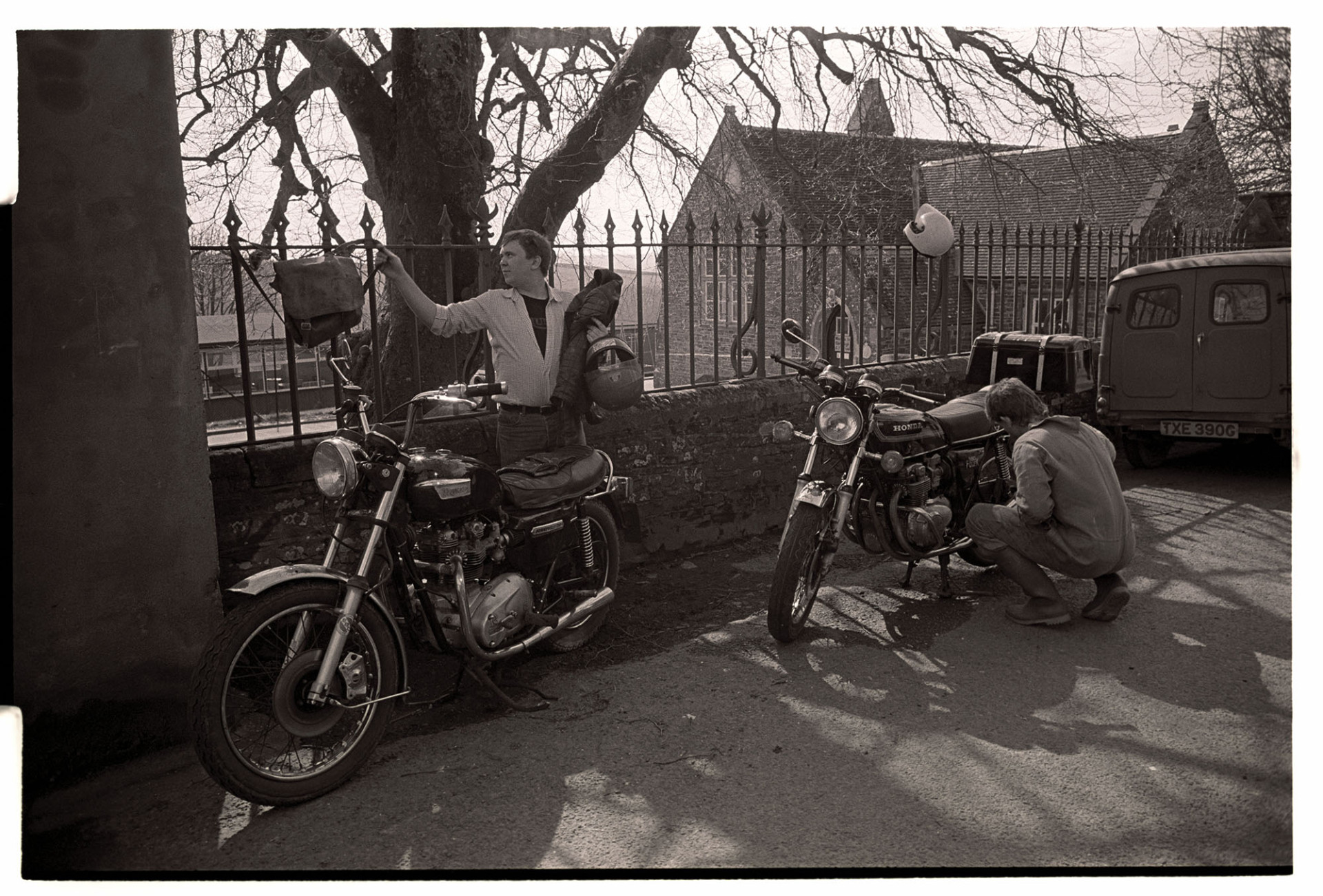 Street scene with parked motorbikes. 
[Two men with their parked motorcycles by railings on a street in Chulmleigh. A building, possibly the school, is in the background.]