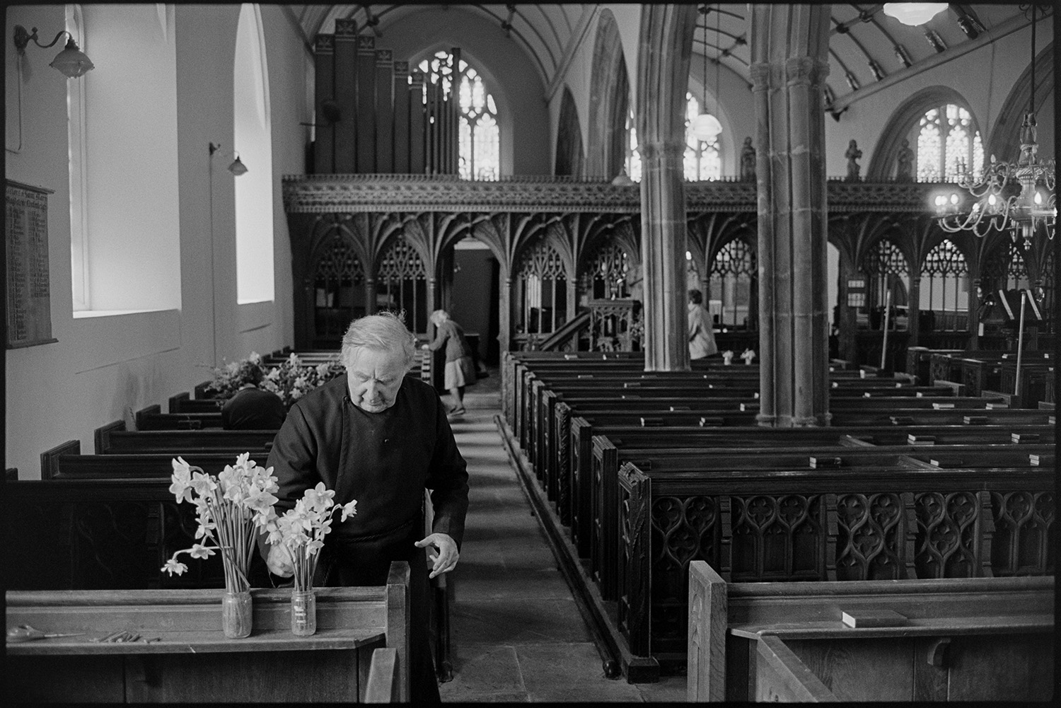 Easter flowers in church. Women chatting and arranging. Fine screen.
[The vicar placing jars of daffodils on a shelf at Chulmleigh Church for the Easter decoration of the church. Carved wooden box pews and a rood screen can be seen in the church.]