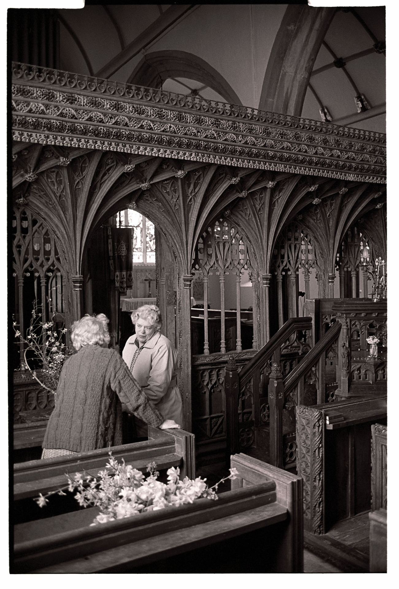 Easter flowers in church, women chatting and arranging. Fine screen. 
[Two women talking and arranging flowers in Chulmleigh Church for Easter. They are stood by the pulpit and rood screen, both of which are ornately carved.]