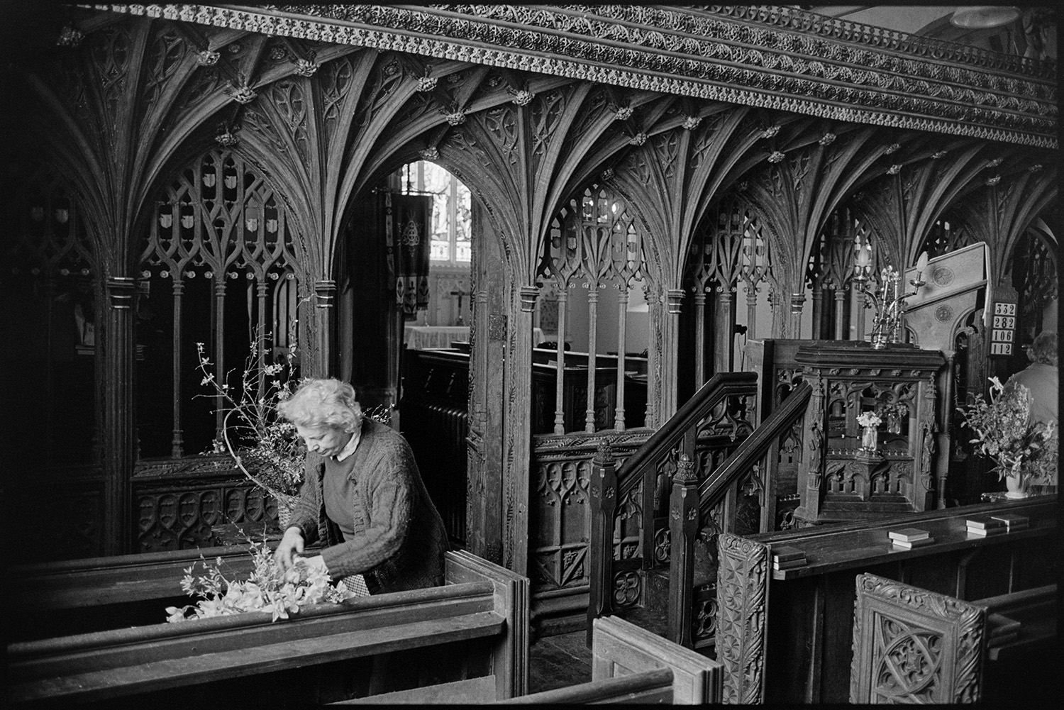 Easter flowers in church. Women chatting and arranging. Fine screen.
[A woman arranging flowers on a pew at Chulmleigh Church for the Easter decoration of the church.  An ornate carved wooden rood screen and steps to the pulpit are visible.]