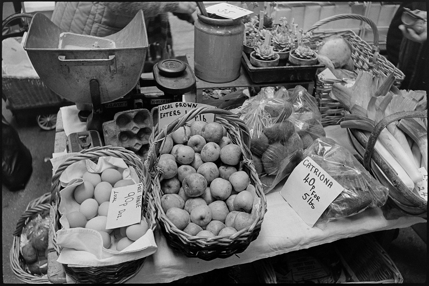 Fruit and vegetable stalls at pannier market, baskets, weighing machine.
[Scales with weights, plants, eggs apples, leeks and other vegetables on a stall in Barnstaple Pannier Market. ]