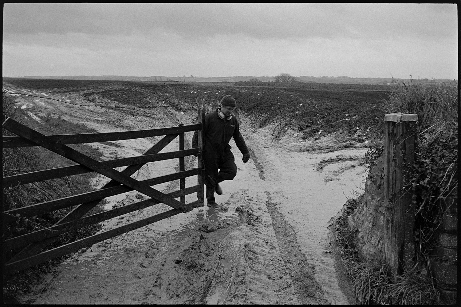 Very muddy field with farmer muckspreading in distance.
[A farmer wearing overalls and ear defenders closing a wooden gate in a very muddy field entrance at Ashreigney.]