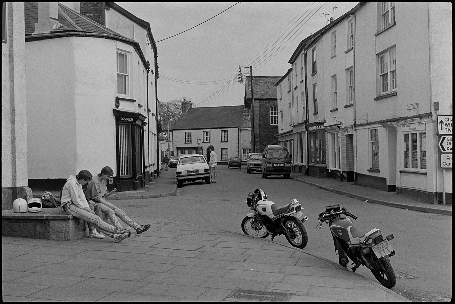 Street scenes with motorbikes.
[Young men sitting on a seat on the side of the street with parked motorbikes and cars in Fore Street, Chulmleigh. Shop fronts can be seen along the street.]