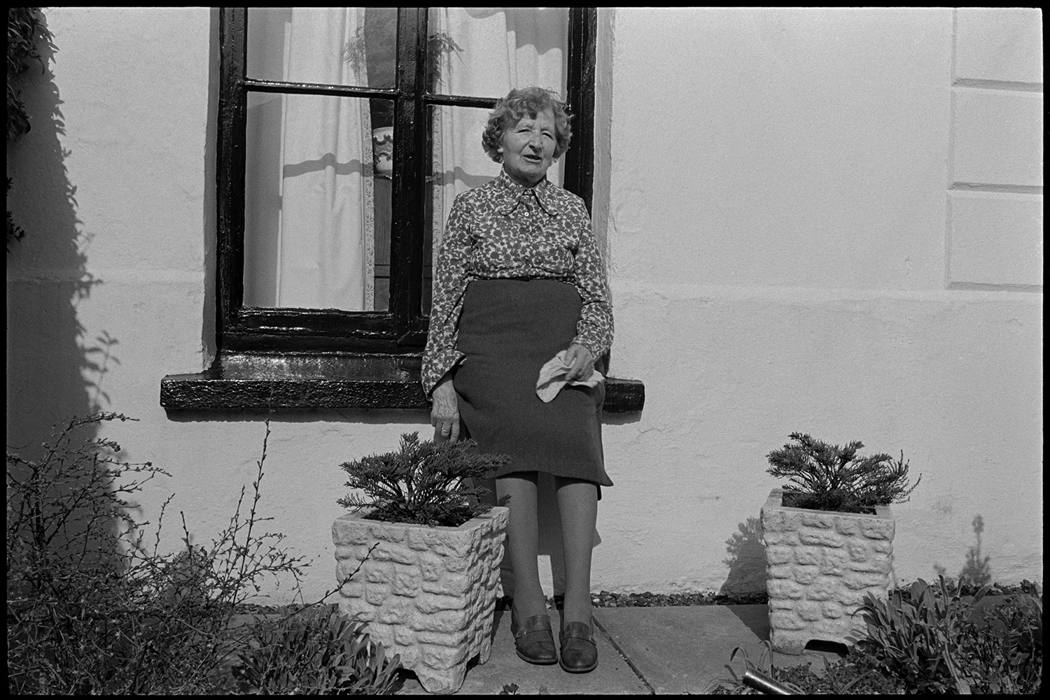 Woman gardening in small patio and sitting chatting to photographer.
[Mrs Bussell sitting on a windowsill in the sunshine in her front garden in Leigh Road, Chulmleigh. Two flower pots are visible in the garden.]