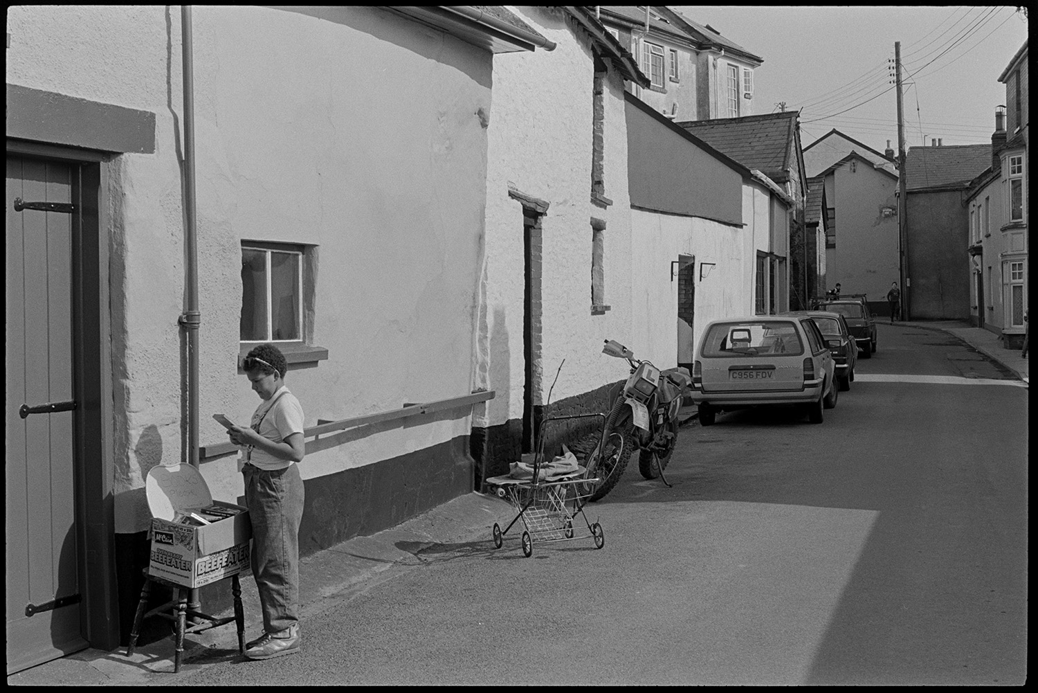 Street scenes with motorbikes.
[A person looking a books in a box on a stool in a street in Chulmleigh. Parked cars, a motorbike and pram wheels are visible along the road.]