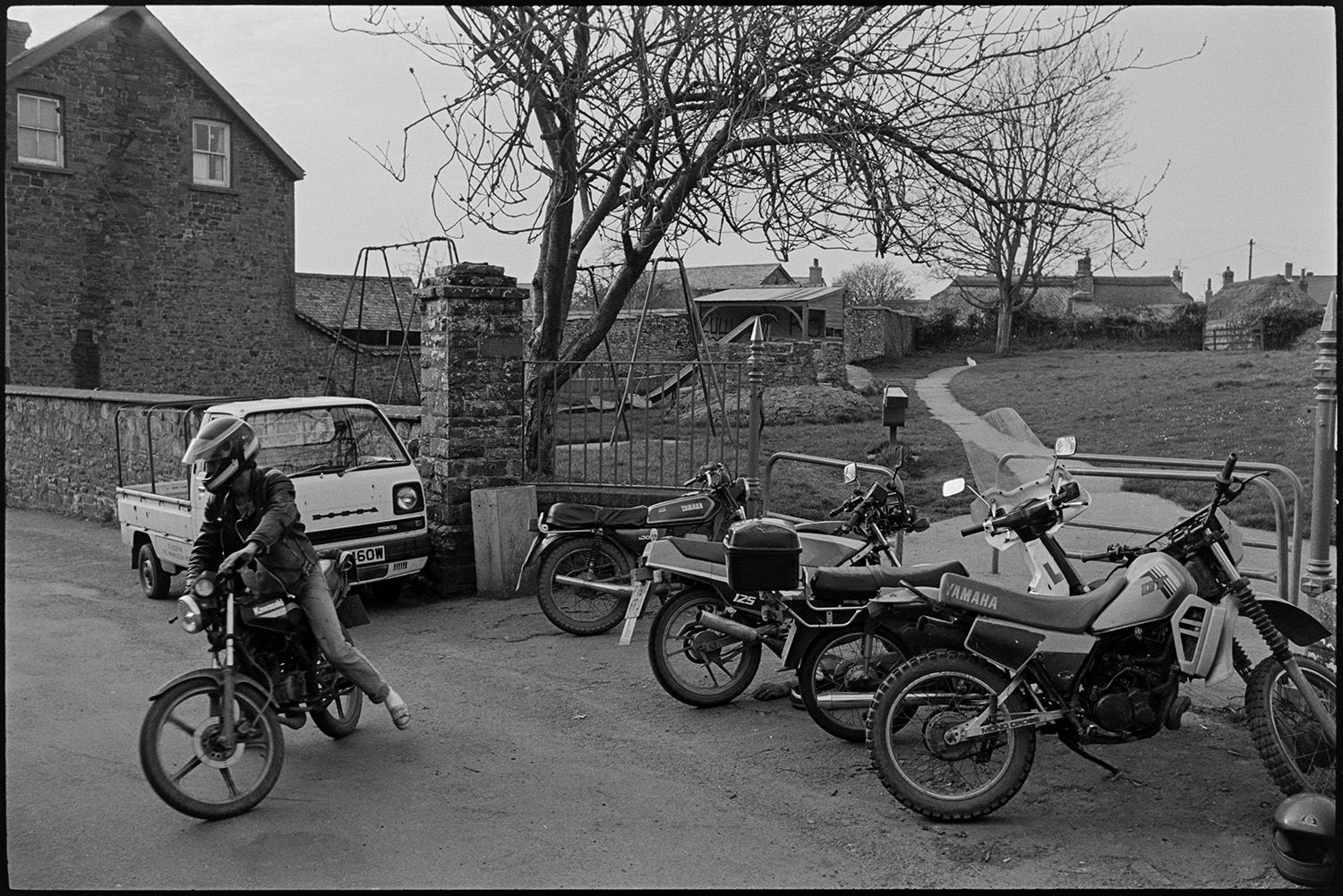 Street scenes with motorbikes.
[Street scene with motorbikes parked at the entrance to the park in Chulmleigh. Swings and a slide can be seen in the park. A person is riding off on a motorbike in the foreground. ]