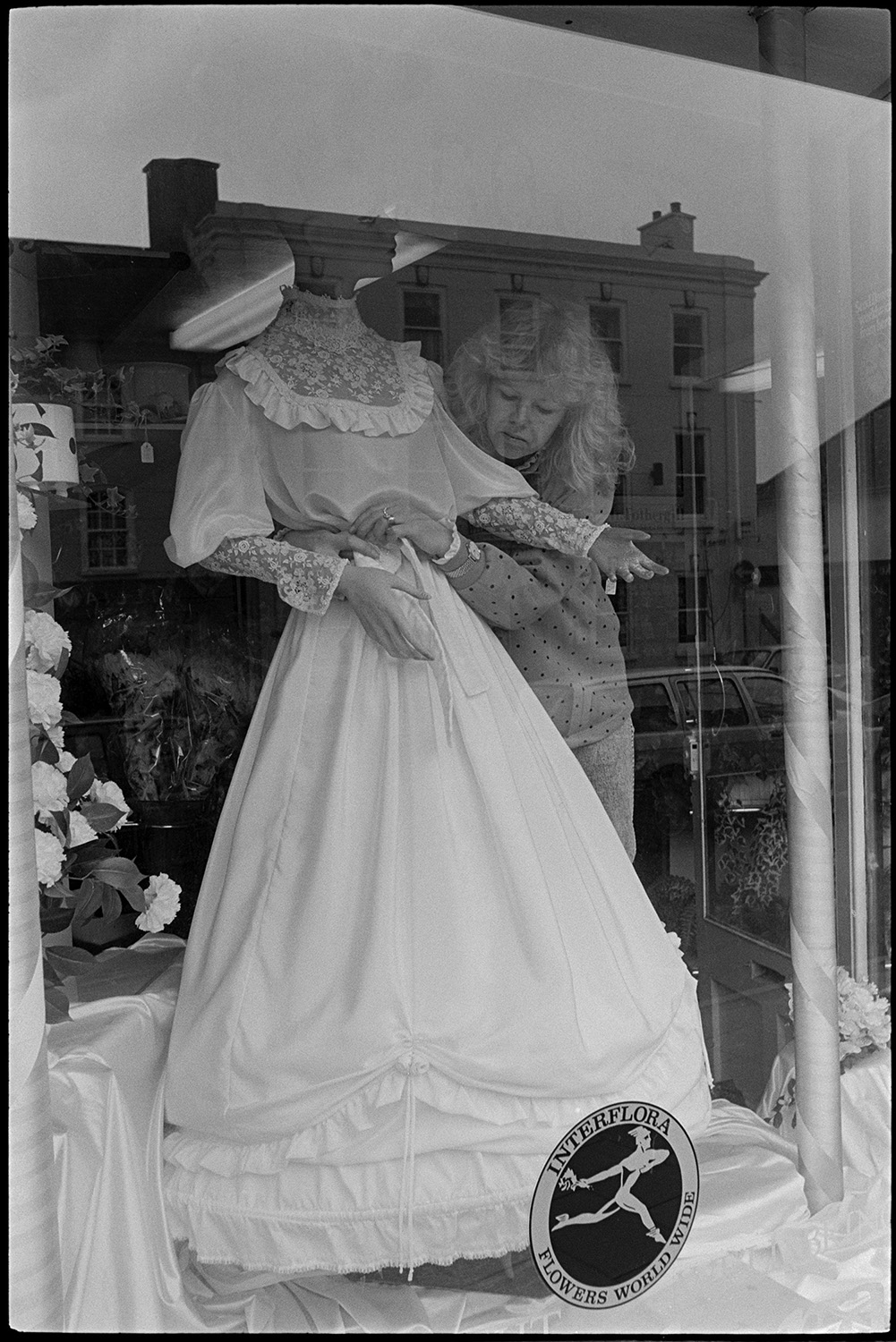 Man and woman arranging Easter wedding display in florists shop window.
[A woman arranging a wedding dress in a florists shop window display for Easter in South Molton.]