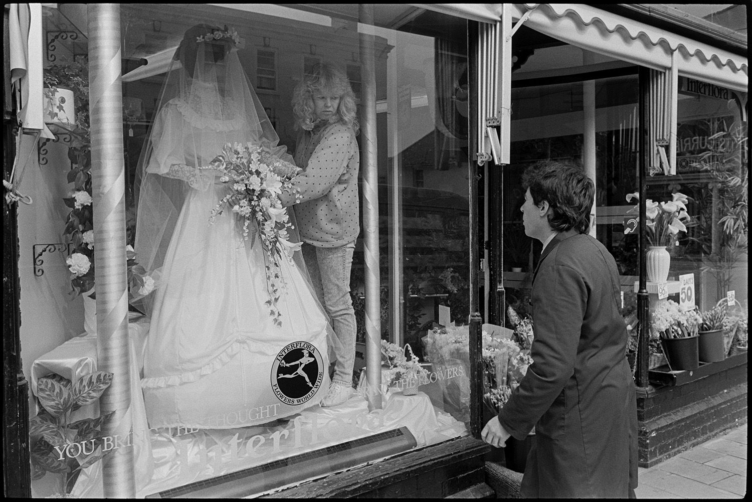 Man and woman arranging Easter wedding display in florists shop window.
[A woman arranging a wedding dress in a florist shop window for an Easter display in South Molton. A man is outside checking the display.]