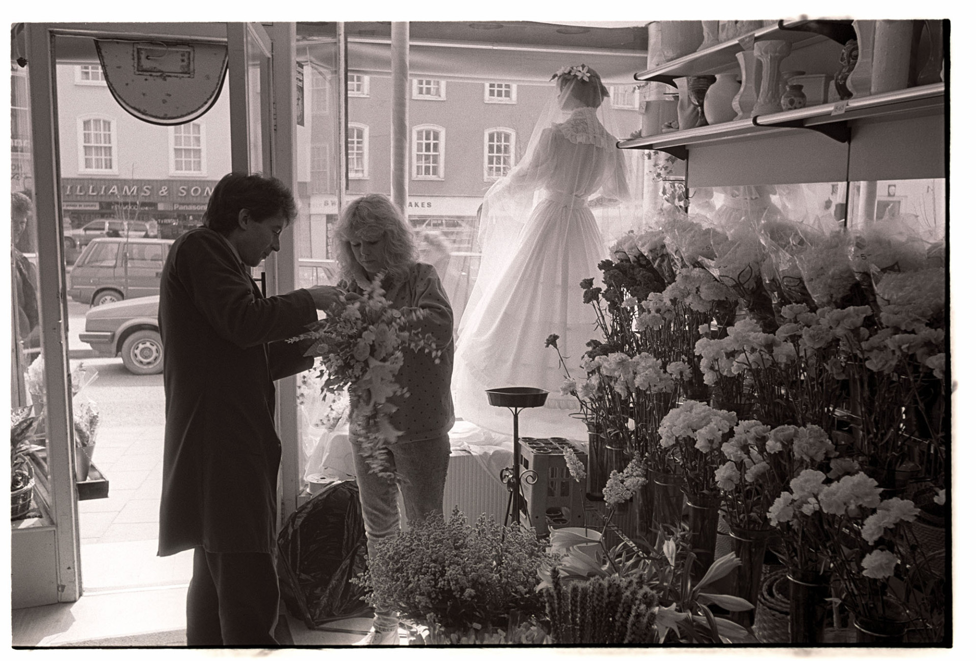 Man and woman arranging Easter wedding display in florists shop window. 
[A man and woman arranging a bouquet in a florists shop in South Molton to put in the shop front window with a mannequin wearing a wedding dress. Flowers and vases are also on display in the shop.]