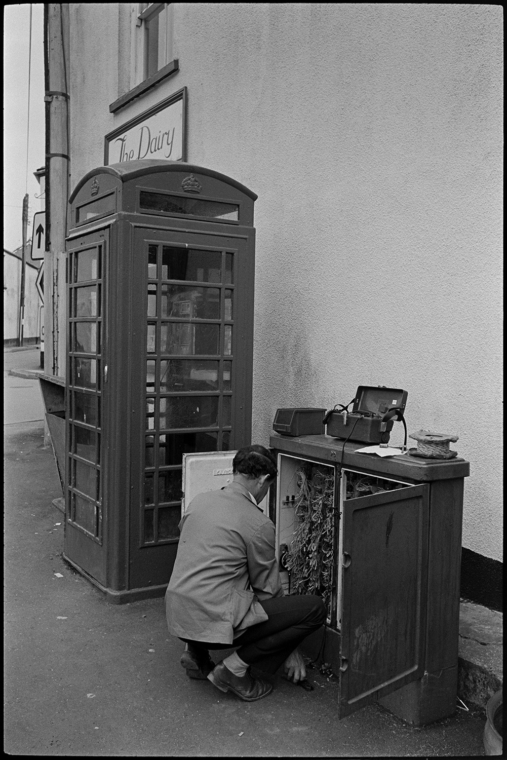 Telephone engineer working in junction box in front of telephone kiosk.
[A man working on telephone wires in a junction box next to a phone box in the street outside The Dairy in Chulmleigh.]