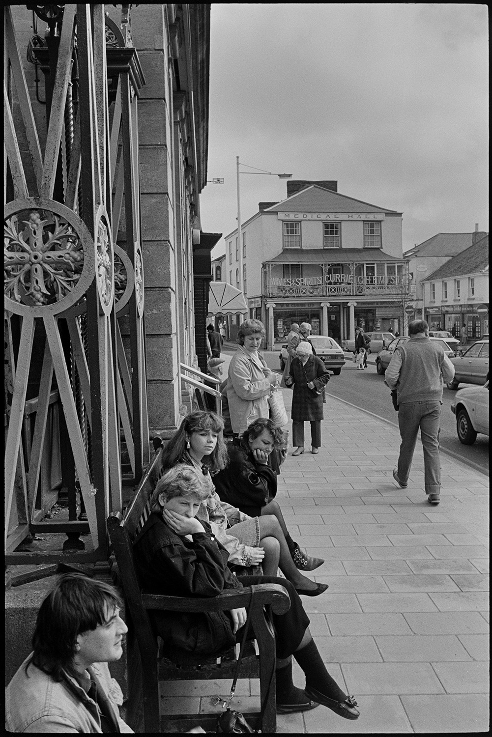 Street scene with people sitting on benches by the wrought iron church gates in South Molton. Currie's chemist shop can be seen in the background beyond cars and passers-by.