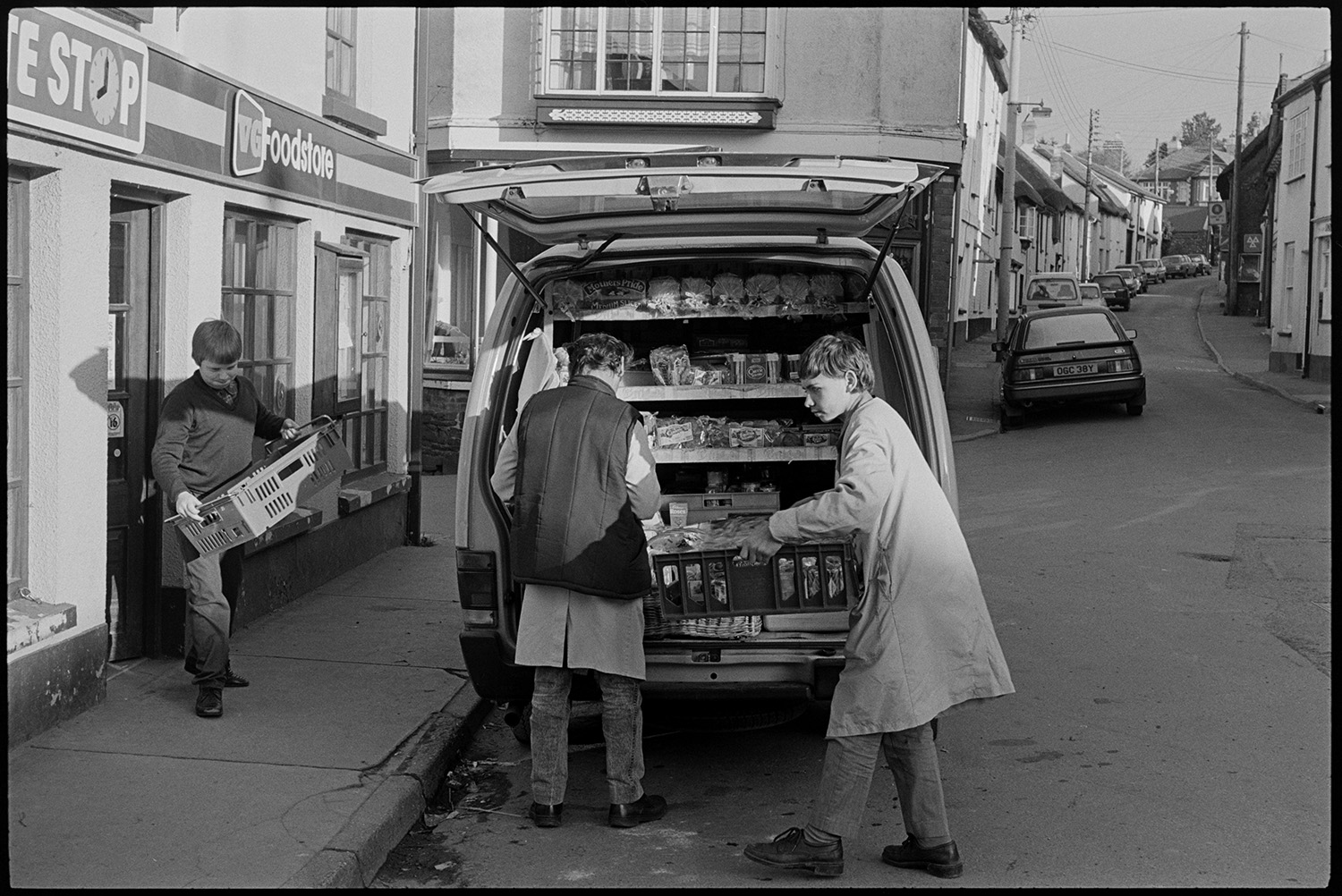Baker making deliveries from van in morning.
[Street scene with people unloading baskets of bread from the back of an open van and carrying it in to the Late Stop shop in Chulmleigh.]