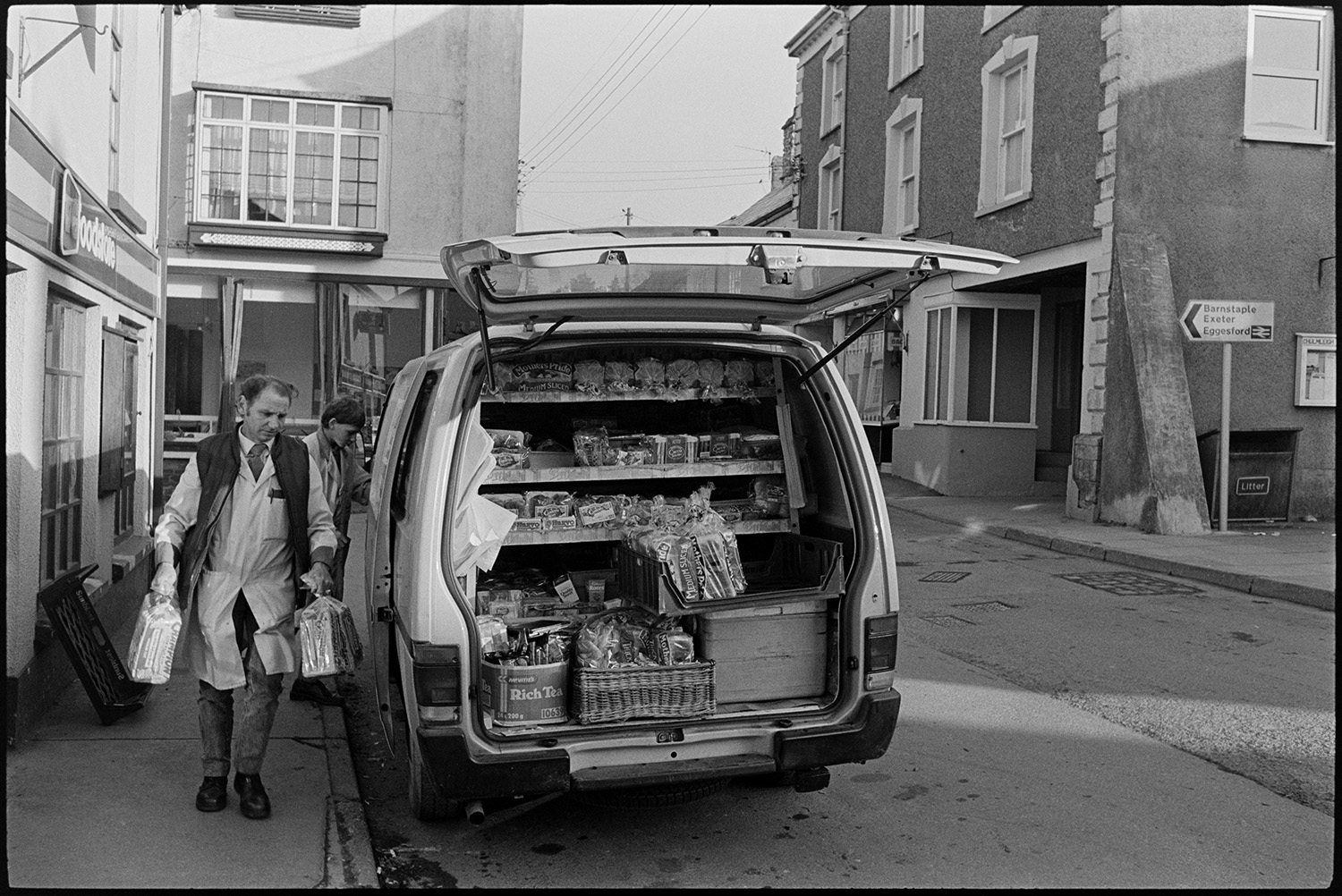 Baker making deliveries from van in morning.
[Street scene with a man unloading bread from the back of an open van in Chulmleigh. Loaves of bread can be seen stacked in the van.]