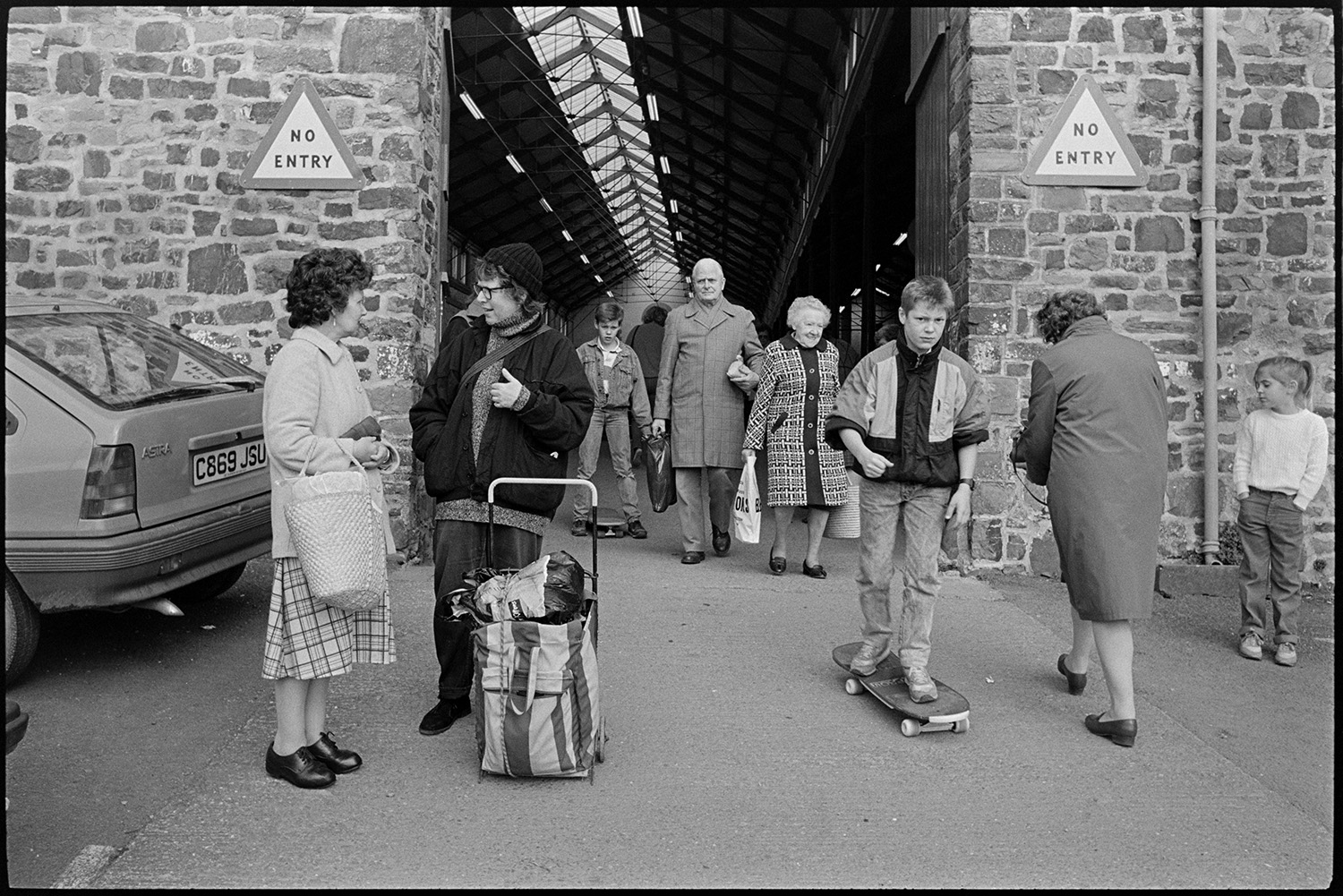 Market hall and cattle market, farmers chatting, loading cows on to trailer.
[People at the entrance to South Molton market, including a boy on a skateboard and two women chatting. There are two no entry signs fixed to the stone wall.]