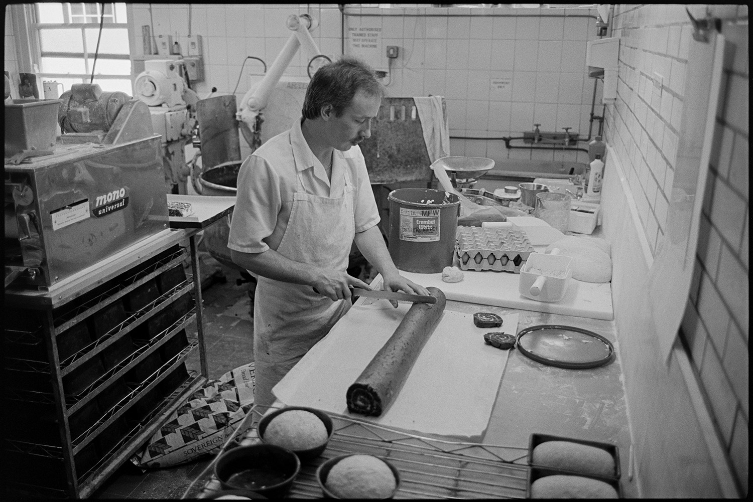 Bakery interior, making bread and serving customers in shop.
[A man working in the white tiled kitchen of The Bakery in East Street, Chulmleigh. He is slicing a chocolate swiss roll on a bench. There is a dough mixing machine along with other ingredients and equipment in the background.]