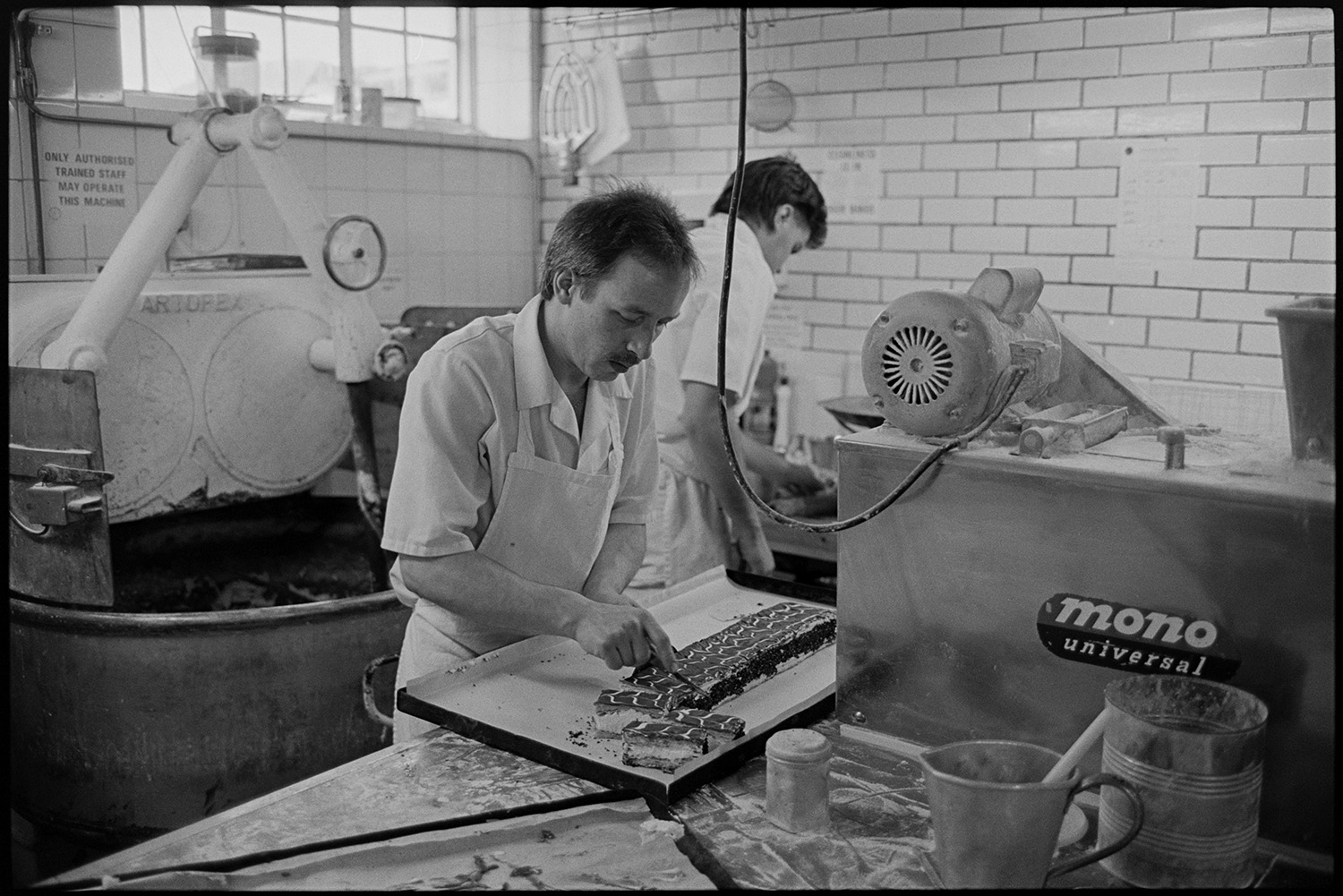 Bakery interior, making bread and serving customers in shop.
[Two men working in the white tiled kitchen at The Bakery in East Street, Chulmleigh. One man is cutting cake slices in a tray on a bench. There is a dough mixing machine along with other bakery equipment in the background.]
