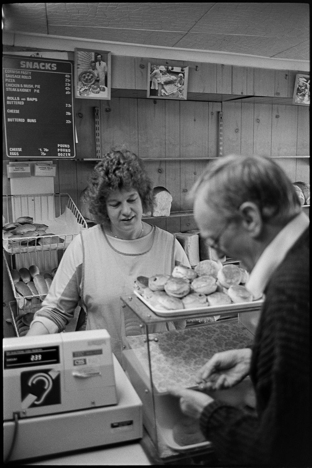 Bakery interior, making bread and serving customers in shop.
[Woman serving a customer in The Bakery shop at East Street, Chulmleigh. There is a tray of buns, shelving with bread and rolls, a till, and a price list on display by the counter. The man in the foreground is counting out money.]