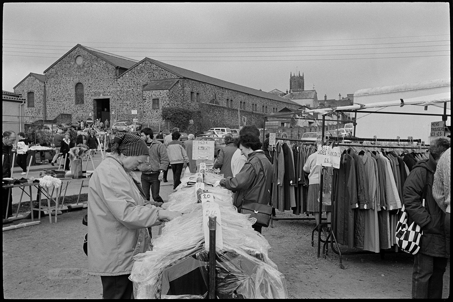 Market hall and cattle market, farmers chatting, loading cows on to trailer.
[People looking around clothes stalls on the site of the South Molton Livestock Market. Two women are looking through a row of coats hanging on a rail. There are rails of clothes, parked cars, stalls, with market buildings and the church tower in the background.]