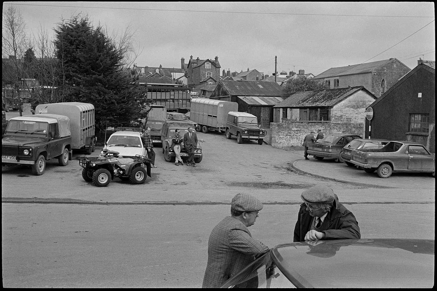 Market hall and cattle market, farmers chatting, loading cows on to trailer.
[Two men talking by a car outside the entrance to the cattle market at South Molton. There are a number of vehicles parked nearby, including quad bikes, jeeps, horse trailers and cattle lorries. Market buildings and houses are visible in the background.]