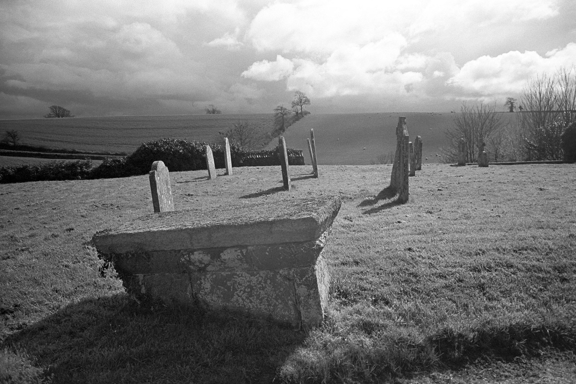 Graveyard with stormy sky, box tomb. 
[A cloudy, stormy sky over Shobrooke churchyard. Gravestone and a box tomb are visible in the churchyard and fields with livestock grazing can be seen in the distance.]
