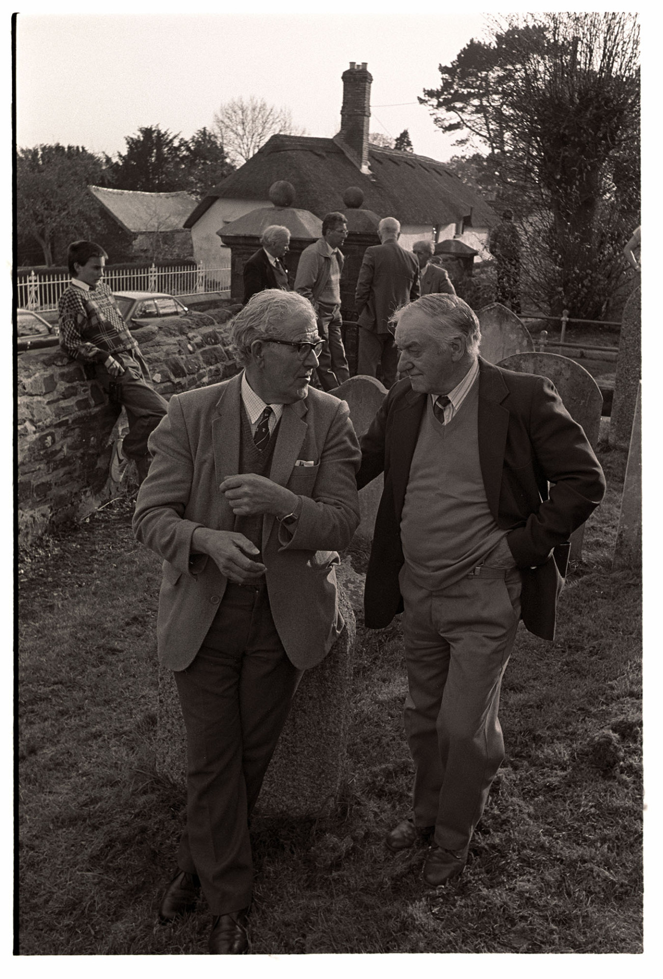 Bellringers standing in graveyard chatting. 
[Bell ringer talking in the churchyard at Chulmleigh at a bell ringing competition. Two men are talking in the foreground, one of which is leaning on a gravestone.]