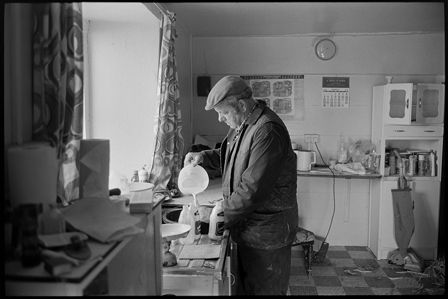 Shepherd feeding lambs beside cob and thatch farmhouse, mixing feed in kitchen.
[John Moyes in the kitchen of Brookland Farm, Chulmleigh filling bottles with milk to feed lambs. Various items can be seen in the kitchen including a hoover, kettle and calendar hung on the wall.]