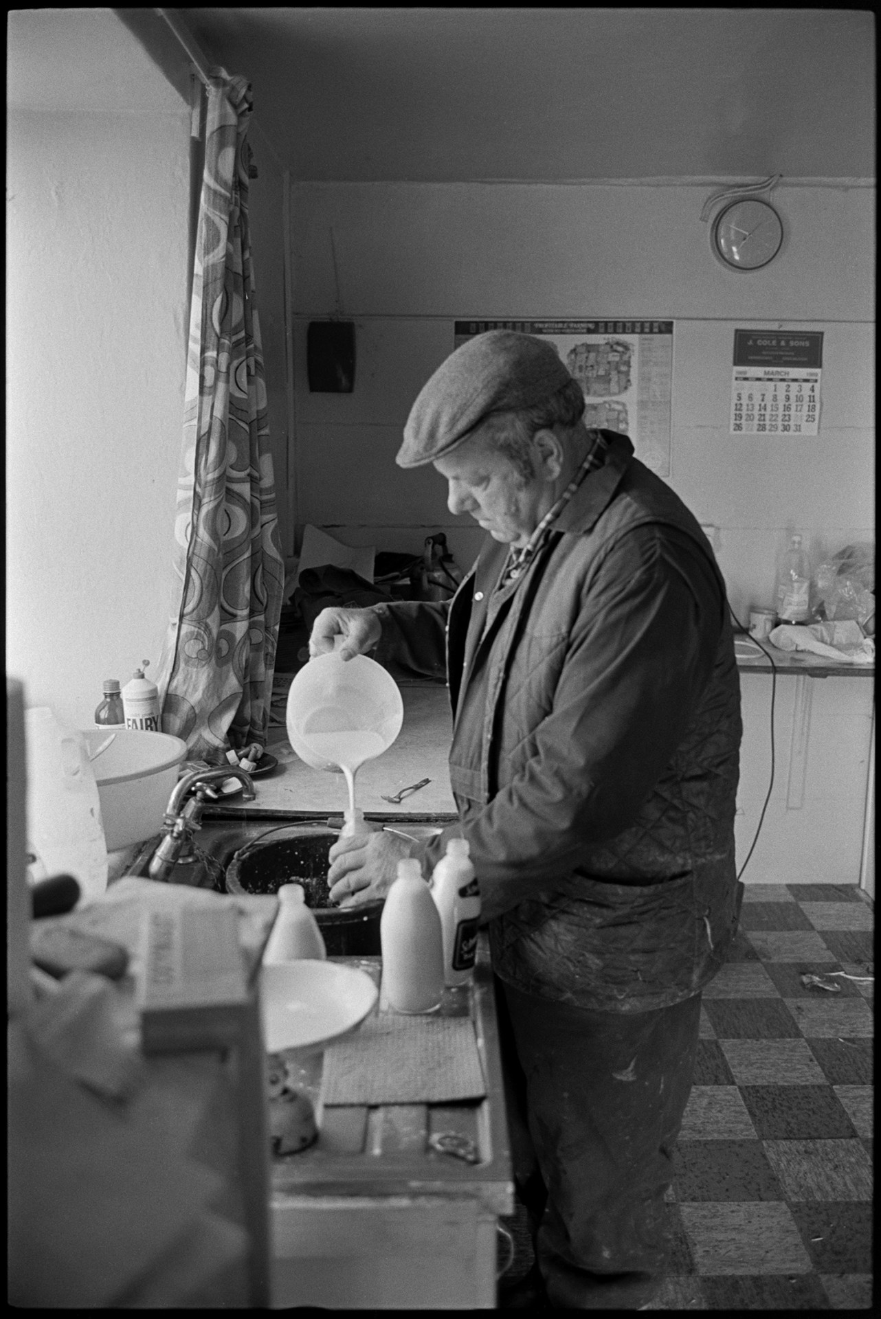 Shepherd feeding lambs beside cob and thatch farmhouse, mixing feed in kitchen.
[John Moyes in the kitchen of Brookland Farm, Chulmleigh filling bottles with milk to feed lambs. Full bottles can be seen on the draining board of the sink.]