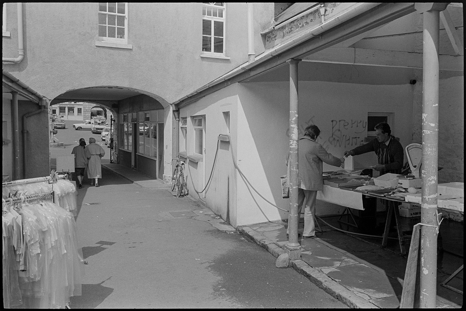 Market row in town.
[A fishmonger serving a customer at a stall in Torrington market row. A set of scales can be seen on the stall and clothes are on display at the shop opposite.  Two women are walking under the entrance arch I the background.]