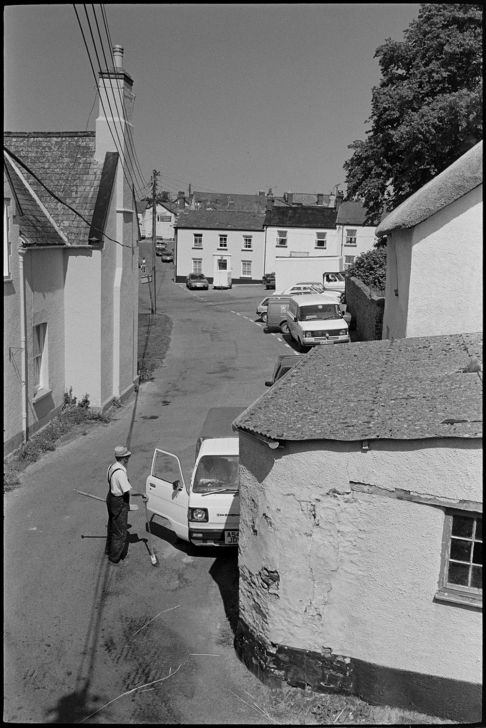 Thatcher working on roof with old building behind, some cob and thatch.
[View of New Street, Chulmleigh, taken from a roof, towards the car parking area in front of the Church of St. Mary Magdalene, Chulmleigh. A man is stood holding a brush and a spade by a parked van in the foreground.]
