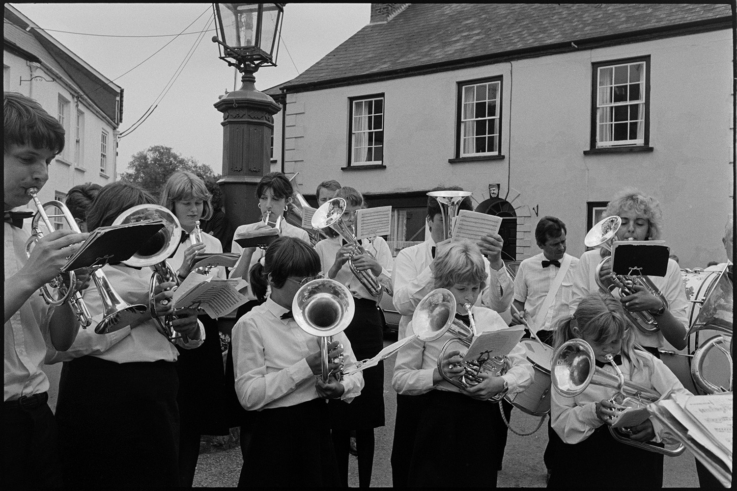 Brass band playing in village.
[South Molton Town Band playing in Fore Street, Chulmleigh at the junction with New Street. The are stood in front of a lamp.]