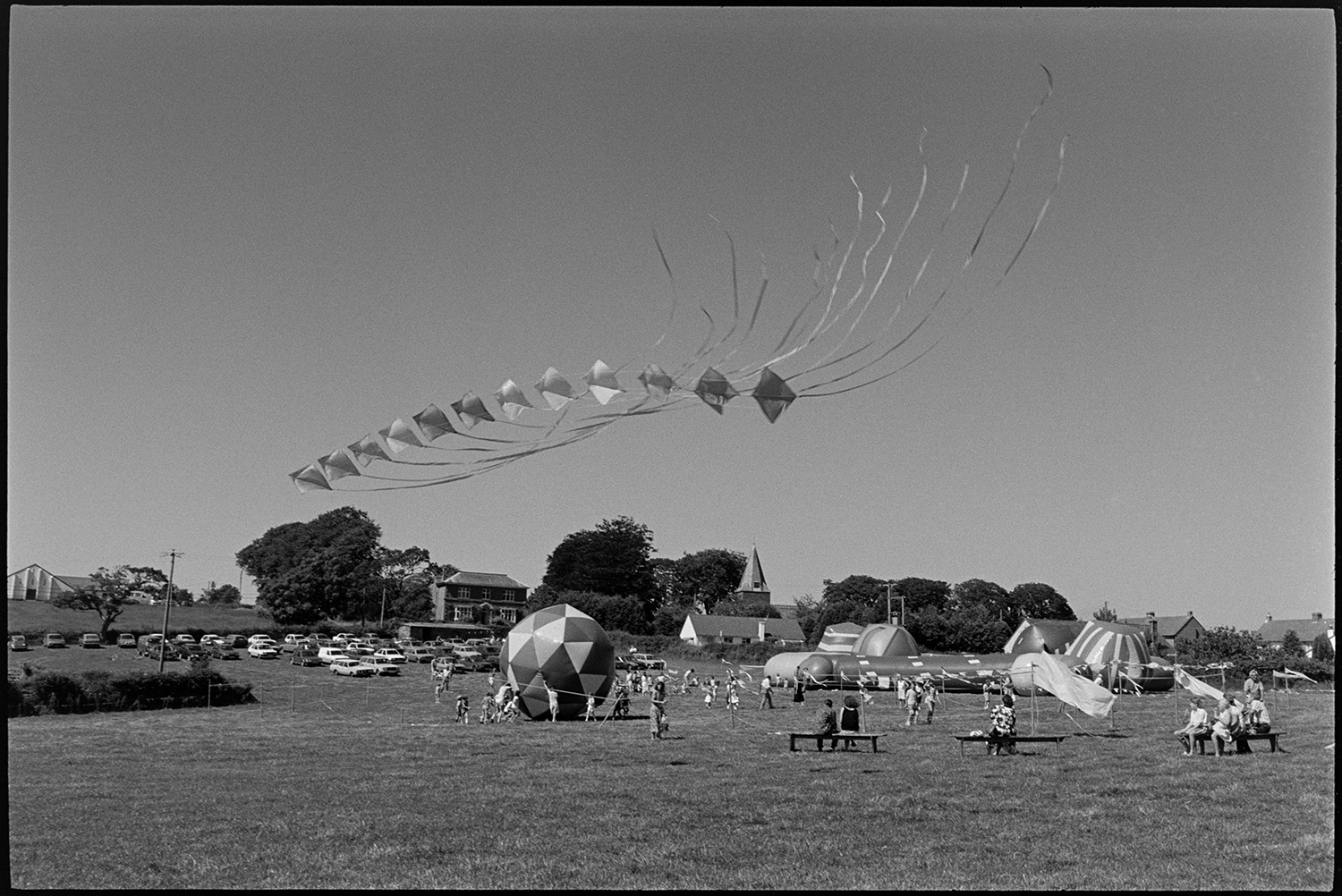 Balloons and kites at Revel.
[Kite display and bouncy castles at the Beaford Revel. Families are sitting on benches watching the display and children are playing. A large number of parked cars are visible in the background.]