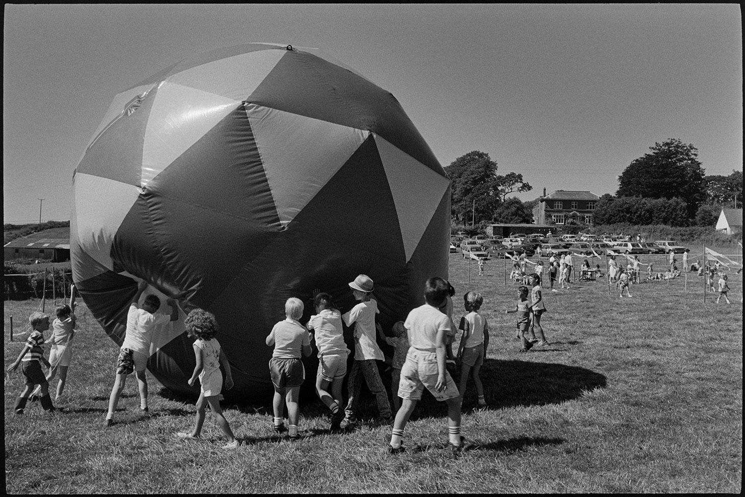 Balloons and kites at Revel.
[A group of children pushing a giant inflated ball at the Beaford Revel. children are playing in the background and a large number of parked cars are visible in the distance.]