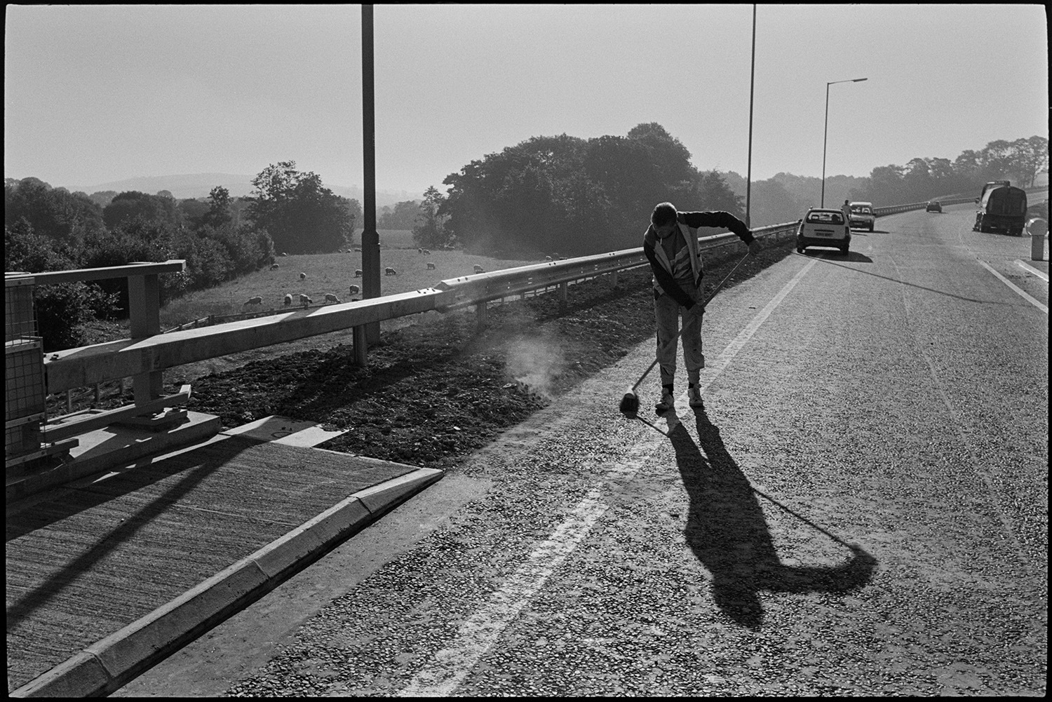 Men sweeping new link road before official opening.
[A man sweeping the A361 North Devon Link Road near South Molton before the official opening. Vehicles ca be seen on the road and in the background and a flock of sheep are grazing in a field adjacent to the road.]