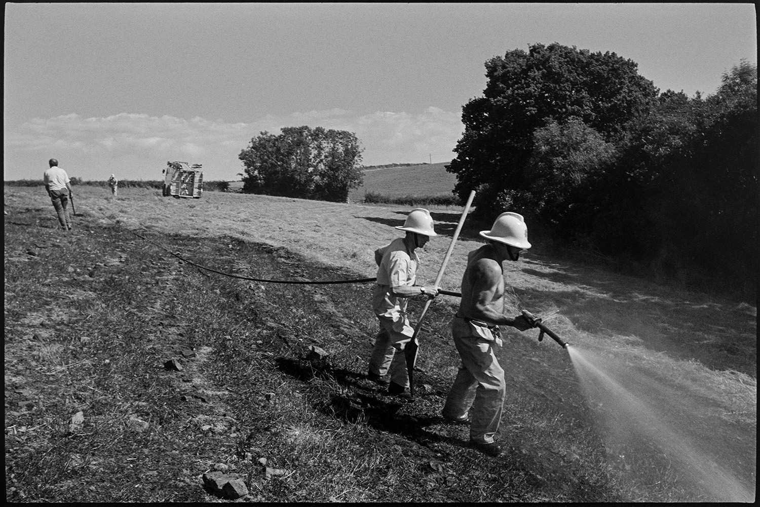Firemen damping down field after crop fire. Fire engine parked in field.
[Two firemen hosing down a crop fire in a field near Chawleigh, with two men and a fire engine in the background. One of the men is holding a beater.]