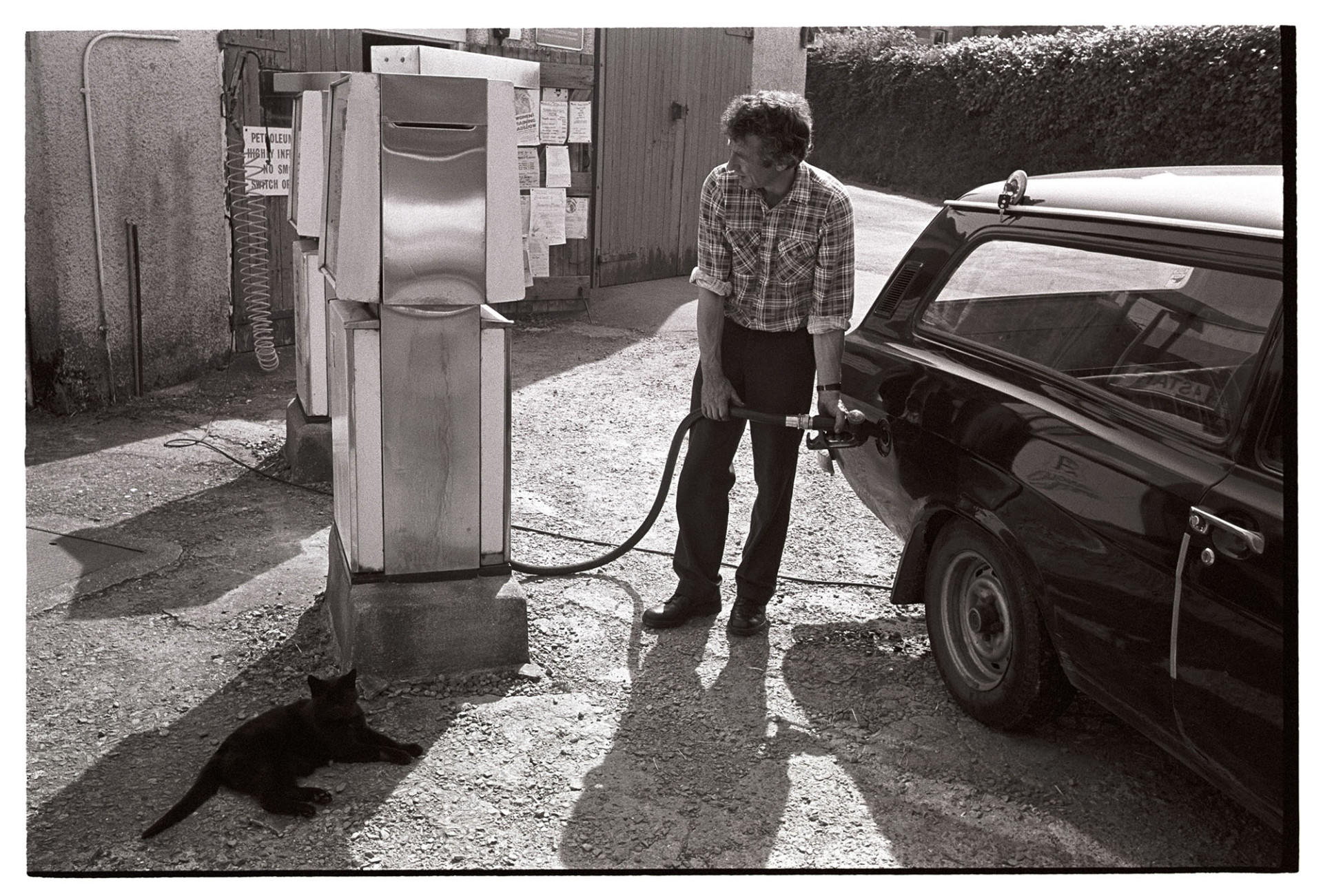 Man filling car with petrol at village garage. 
[A man filling his car with petrol from a petrol pump at the village garage in George Nympton. A cat is sat by the petrol pump.]