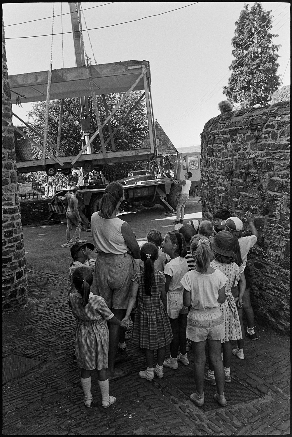 Children watching crane remove portacabin school building.
[A group of children and a woman watching two men manoeuvring the frame of a portacabin being hoisted by crane on to a lorry. The portacabin was previously used as a school building.]