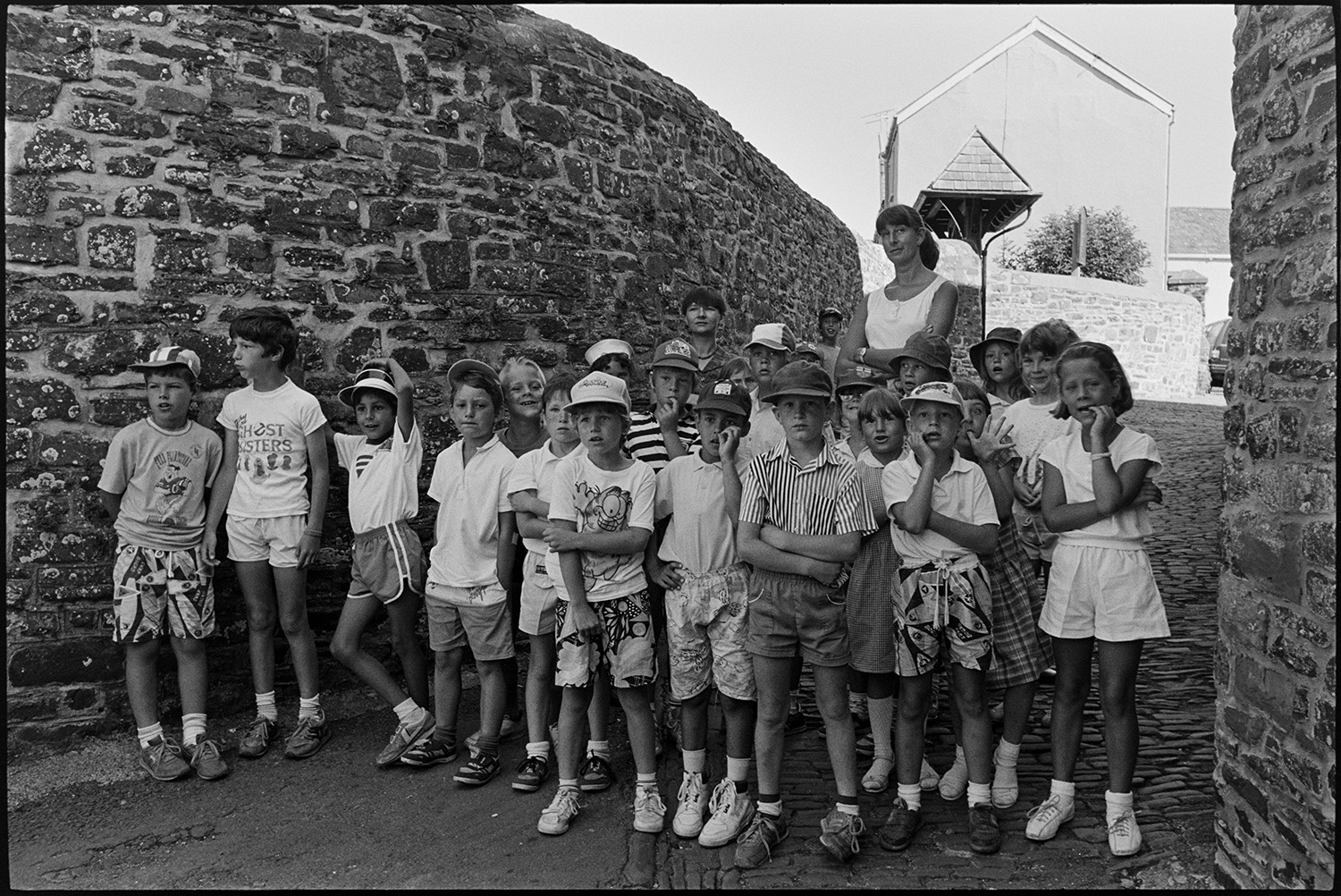 Children watching crane remove portacabin school building.
[A group of children and a woman watching the removal of a portacabin previously used as a classroom at Chulmleigh Primary School. They are stood on a cobbled pathway between two walls.]