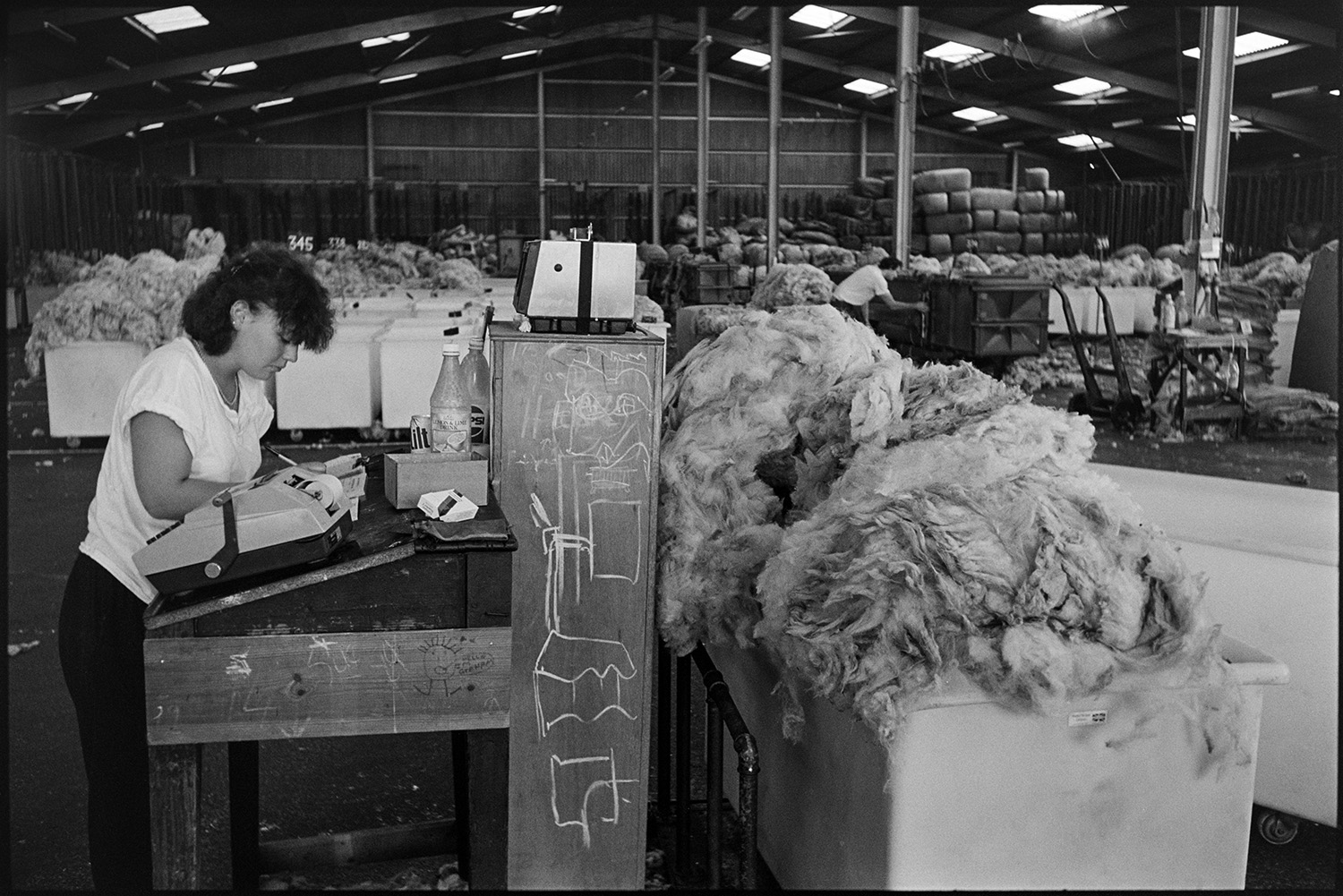 A woman recording information next to a receipt machine at a desk in the warehouse of Devon & Cornwall Wools Ltd at the Pathfields Business Park in South Molton. She is surrounded by bins of wool and fleeces.