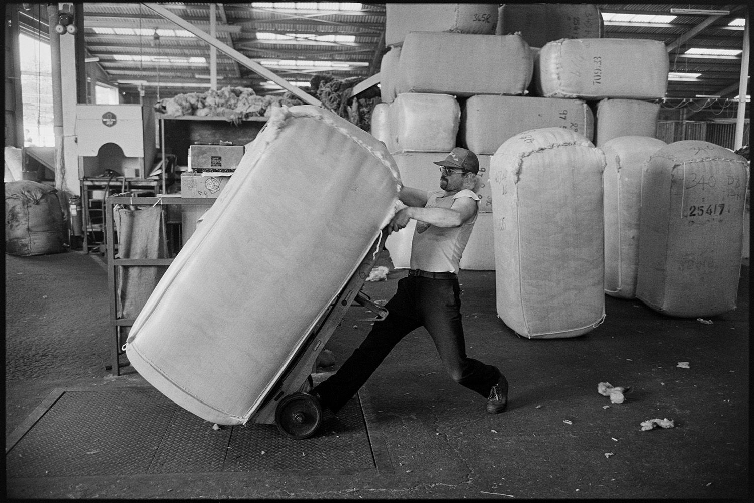 Sorting and packing wool, stacks of bales, office with paper work.
[A man wheeling a large bale of wool on a trolley or sack truck in the warehouse of Devon & Cornwall Wools Ltd at the Pathfields Business Park, in South Molton, with stacks of bales behind him.]