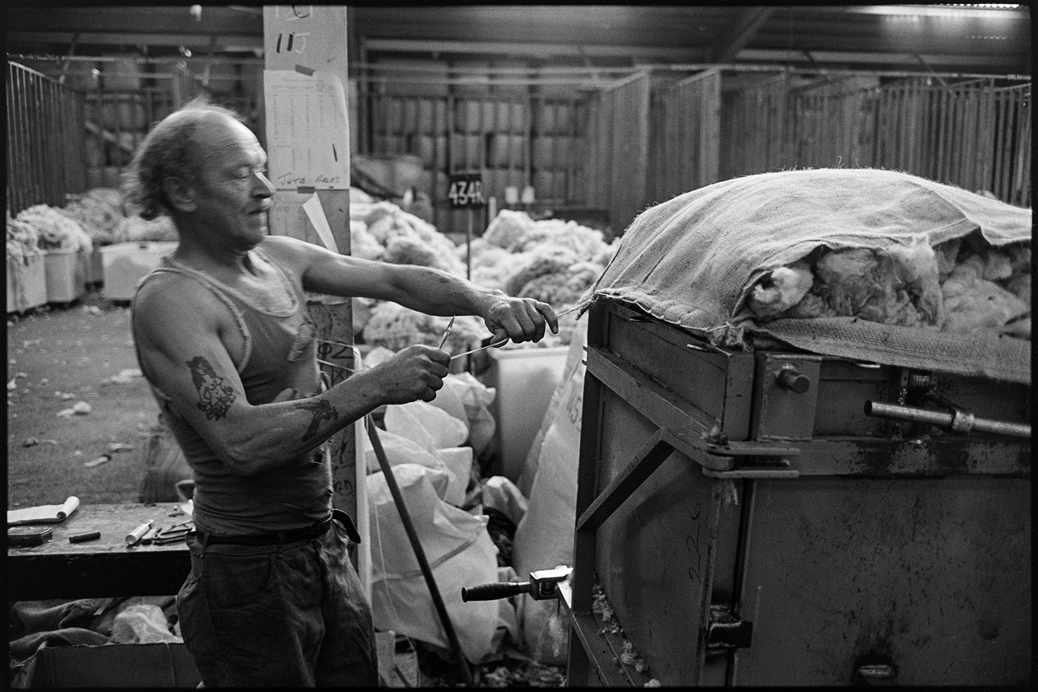 Sorting and packing wool, stacks of bales, office with paper work.
[A man with tattooed arms fixing a cover over the top of a container of wool in the warehouse of Devon & Cornwall Wools Ltd at the Pathfields Business Park in South Molton.]