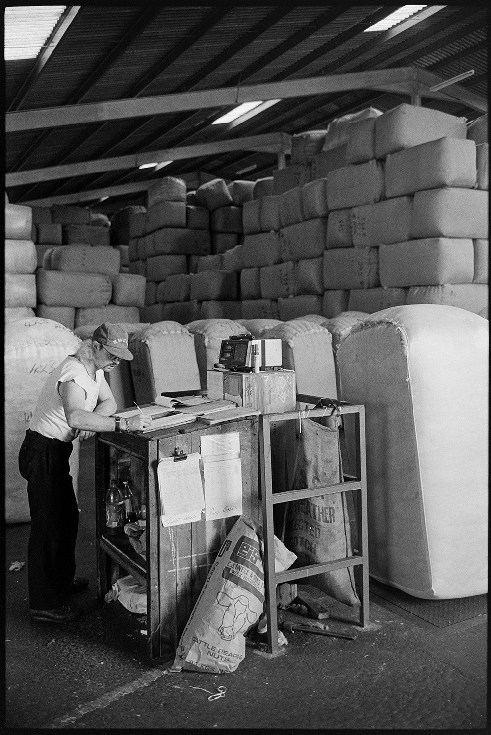 Sorting and packing wool, stacks of bales, office with paper work.
[A man recording information at a desk in the warehouse of Devon & Cornwall Wools Ltd at the Pathfields Business Park in South Molton. He is surrounded by stacked bales of wool.]