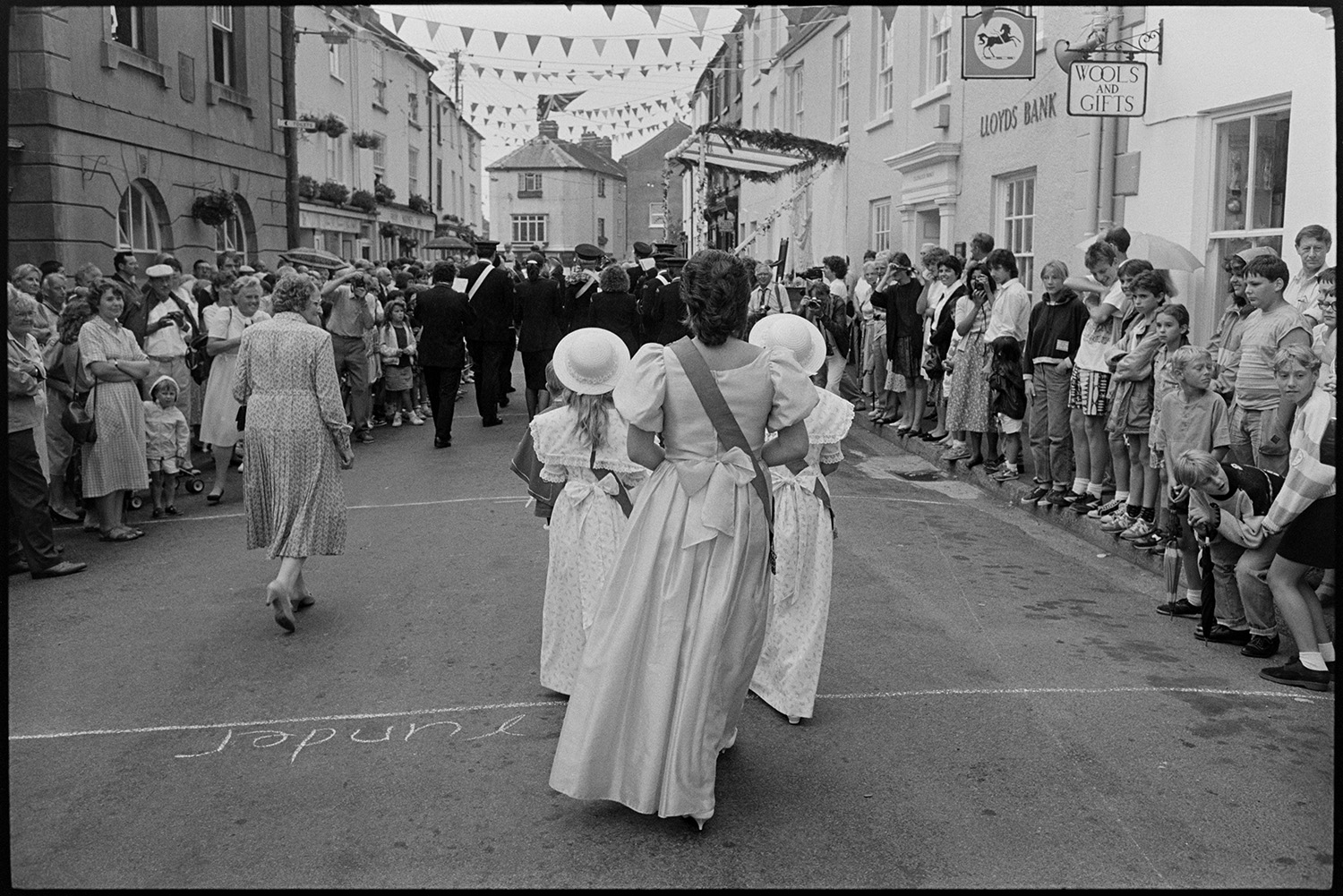 Chulmleigh Fair, Queen and attendants leaving church and processing through village.
[The Chulmleigh Fair Queen and attendants processing along Fore Street, Chulmleigh behind a marching band at Chulmleigh Fair. People are watching from both sides of the street. The street is decorated with bunting and Lloyds Bank can be seen and a sign for 