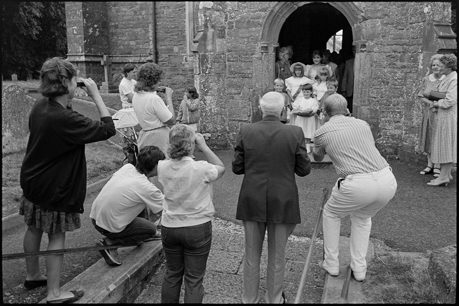 Chulmleigh Fair, Queen and attendants leaving church and processing through village.
[People taking photographs of the Chulmleigh Fair Queen and her attendants stood in the porch of the Church of St Mary Magdalene, Chulmleigh. before parading down to the town centre for Chulmleigh Fair.]]