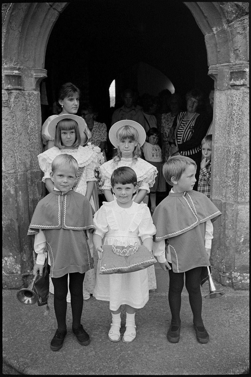 Chulmleigh Fair, Queen and attendants leaving church and processing through village.
[Boys and girls dressed for their role as attendants to the Chulmleigh Fair Queen standing in front of the entrance to the Church of St Mary Magdalene, Chulmleigh. The two boys are carrying trumpets and one of the girls is carrying a crown resting on a cushion. The Fair Queen and other women are gathered in the background.]