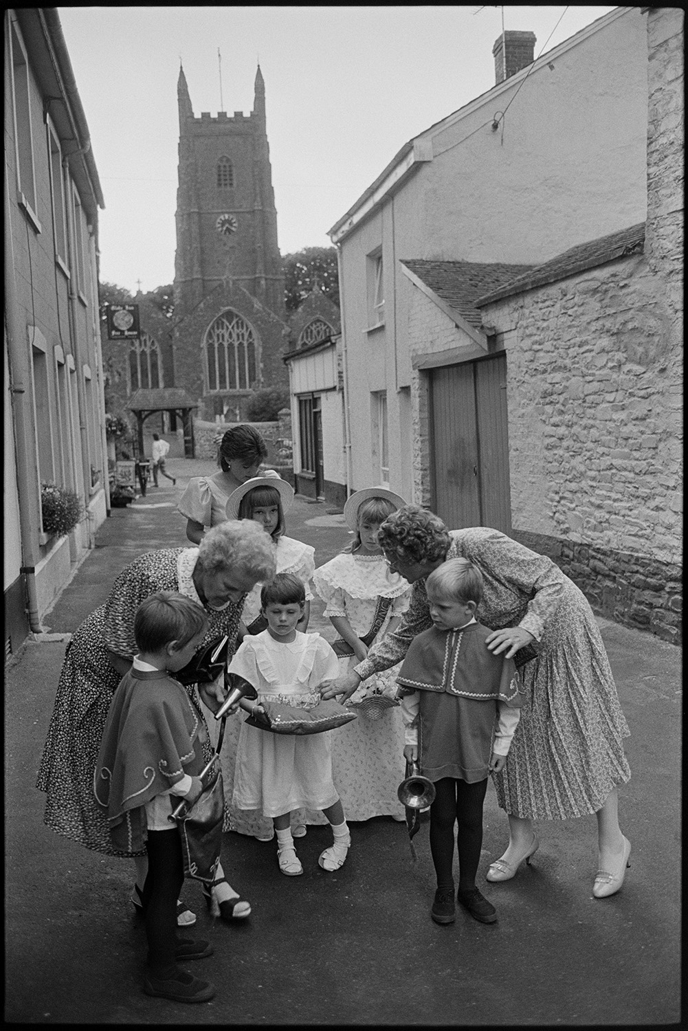 Chulmleigh Fair, Queen and attendants leaving church and processing through village.
[Two women helping a group of young children who are acting as attendants to the Chulmleigh Fair Queen who is standing behind them, in New Street, Chulmleigh. The Church of St Mary Magdalene is visible in the background. The boys are holding trumpets and the girl in the foreground is holding a crown resting on a cushion.]