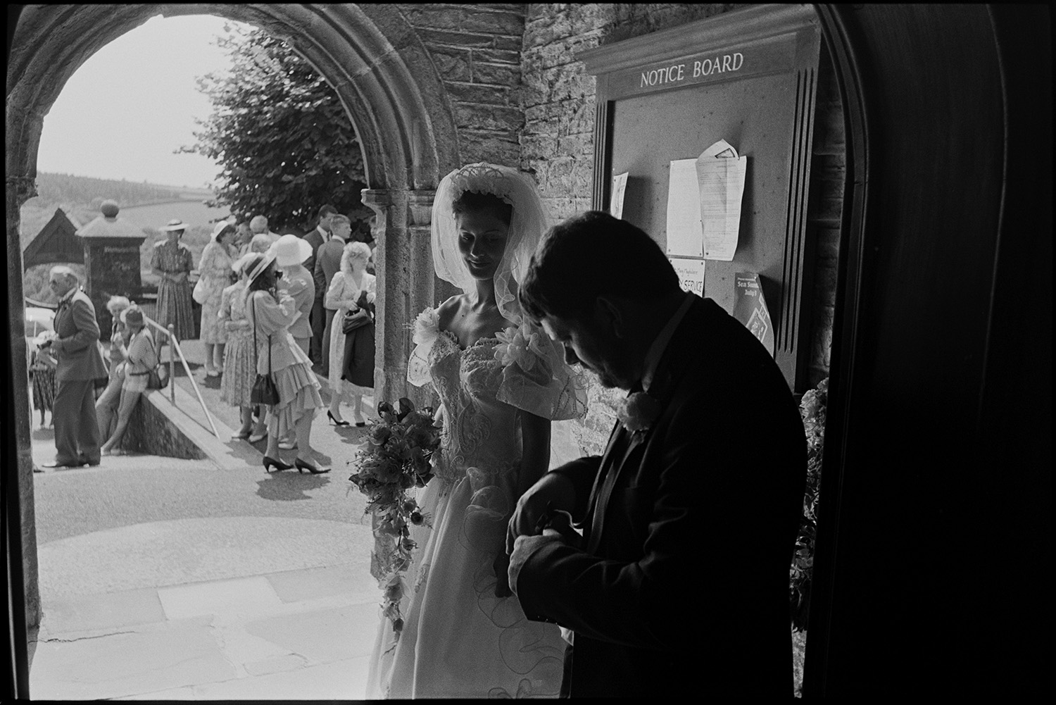 Wedding spectators, bride, groom, bridesmaids being photographed Photographer changing film.
[A bride and man standing in the porch of the Church of St Mary Magdalene, Chulmleigh. The bride is holding a bouquet of flowers and watching the man changing a film in his camera. Guests can be seen waiting outside the church.]